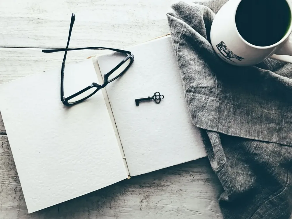 Looking down at an empty page in a notebook with key and glasses on top which are layin gon a wooden table
                with a blanket next to it with a cup of coffee on top.